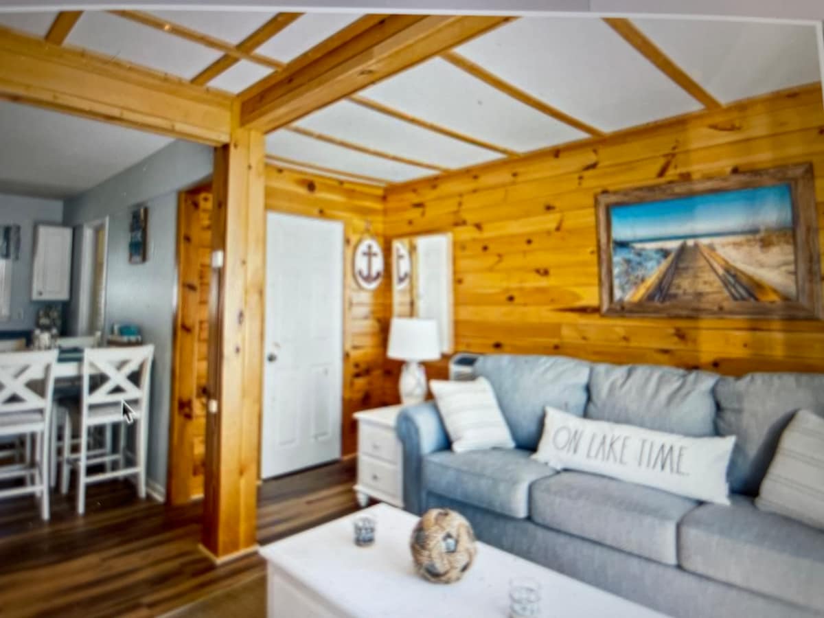 A living area is presented with a light blue sofa adorned with decorative pillows. Natural wooden walls feature nautical-themed artwork and a wall clock. A white coffee table sits in front of the sofa, while a dining area with high chairs is visible in the background.