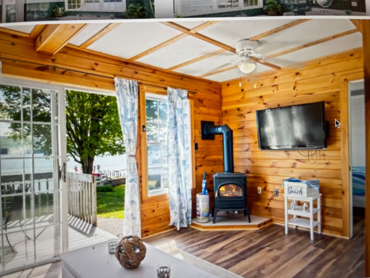 A bright living area features wooden walls and a rustic ceiling with exposed beams. A cozy wood stove is positioned beside a television on the wall. Natural light enters through large windows adorned with light blue curtains, providing views of the outdoor setting.