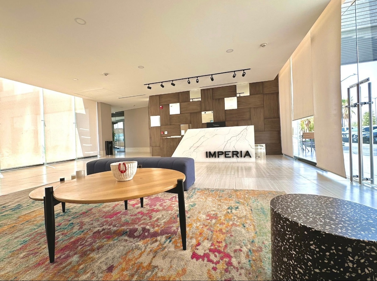 A spacious lobby is highlighted by a sleek reception desk with the name 'IMPERIA' prominently displayed. Modern furniture includes a low wooden coffee table and a comfortable dark sofa, all set on a vibrant area rug. Natural light illuminates the area through large glass windows.