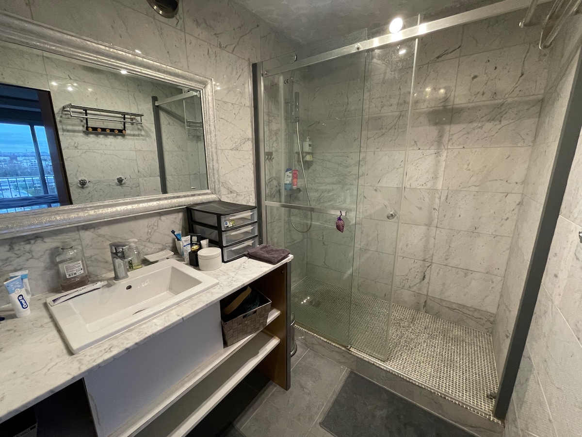 The bathroom features a large glass shower enclosed by a transparent wall, complemented by marble tiles throughout. A modern vanity with a sink is located below a mirror, and several toiletries are neatly arranged on the countertop. Natural light enters through a nearby window.