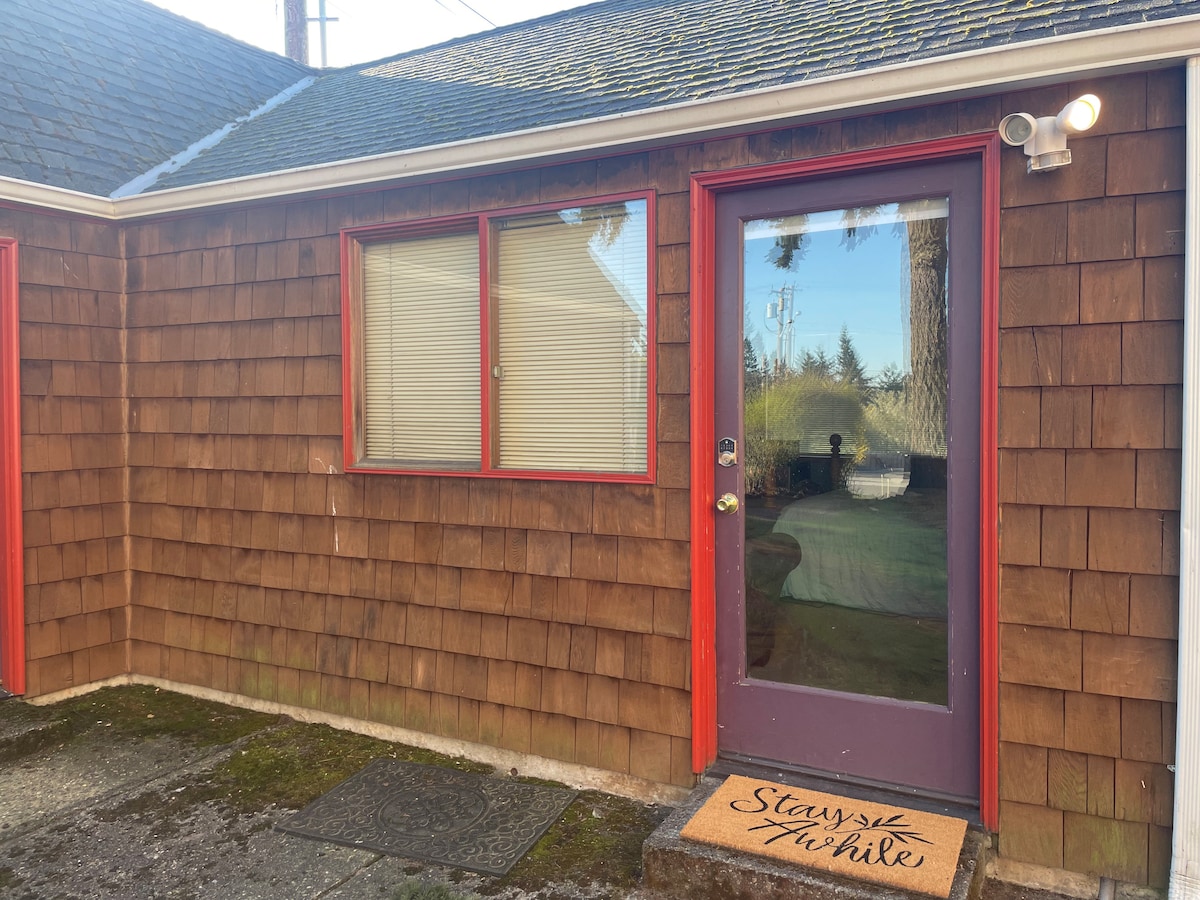 The entrance features a door framed in vibrant red, leading into the guest house. A large window allows natural light to filter in, and a welcome mat is situated at the entrance. The exterior is clad in deep brown shingles, complemented by a mossy stone pathway.