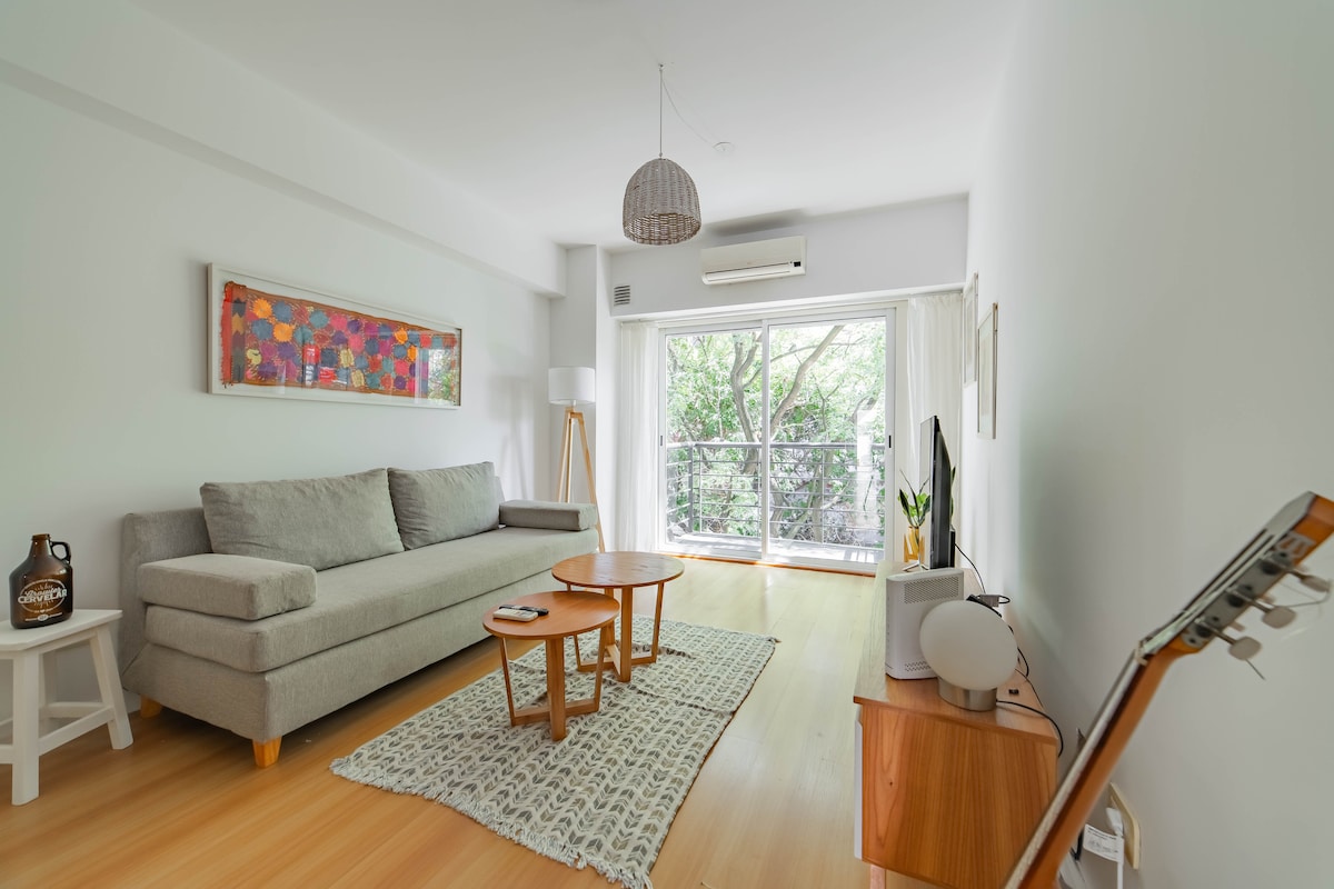 The living area features a comfortable sofa and two wooden tables positioned on a light-colored floor. A large window allows natural light to illuminate the space, with a view of greenery visible outside. A guitar rests against a wooden cabinet, adding a homely touch.
