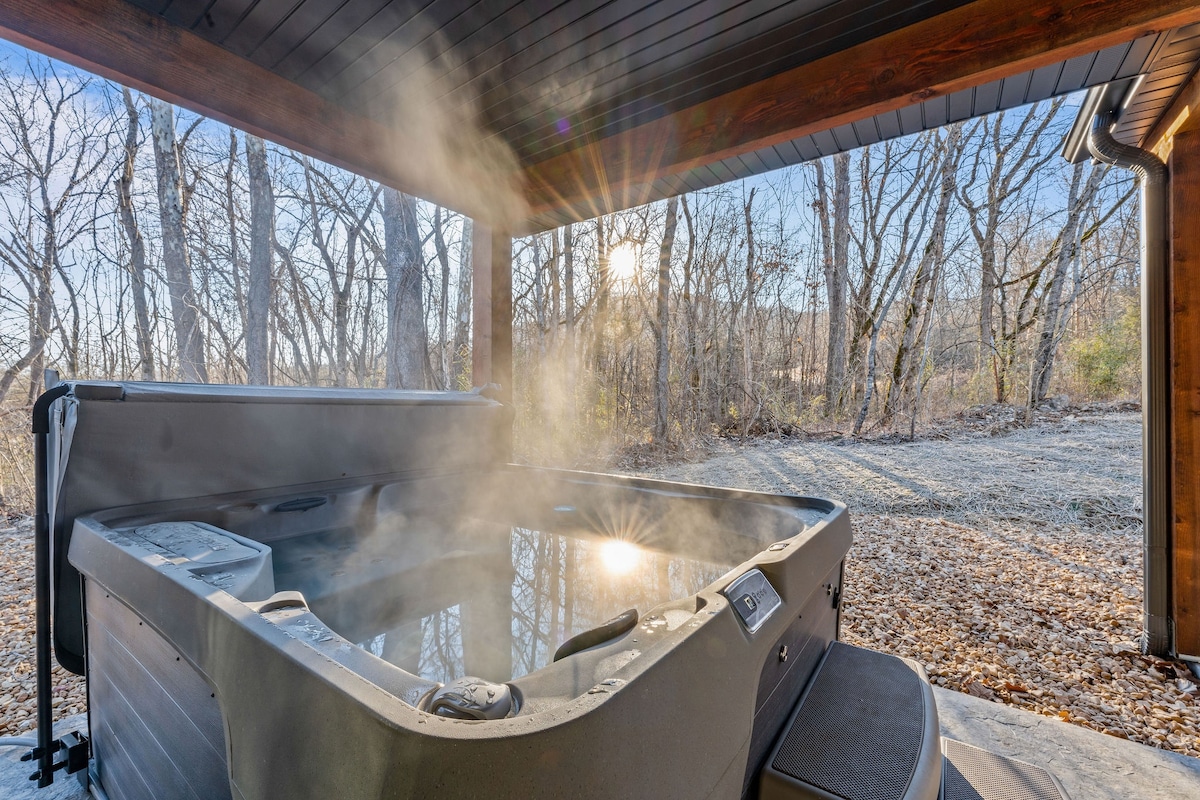 A hot tub is featured on a covered patio, with steam rising in the fresh air. Surrounding trees create a natural backdrop, while sunlight filters through, enhancing the serene atmosphere of the outdoor space.
