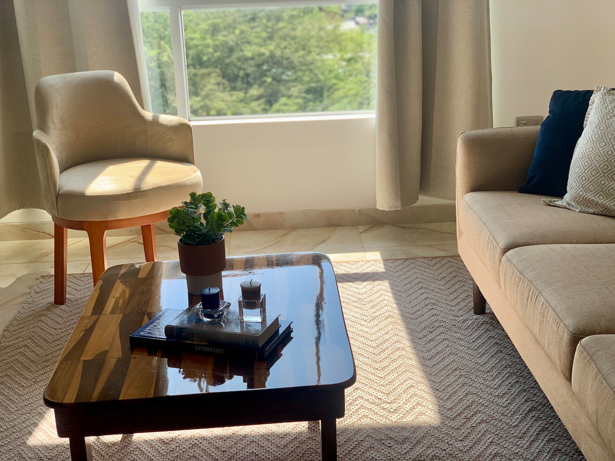 A bright living area features a comfortable sofa and an armchair positioned near a large window that offers views of greenery. A coffee table with a plant and easily accessible books enhances the inviting space, while soft curtains frame the light-filled room.