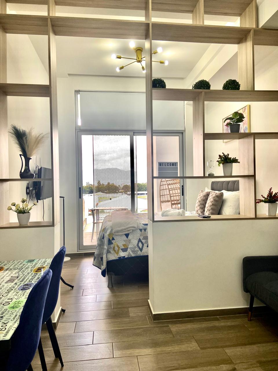 A bright living space is framed by modern shelving, displaying decorative plants and chic accessories. A spacious bed is visible through a glass door that leads to a balcony, while a small dining area is arranged with stylish chairs and a patterned tablecloth.