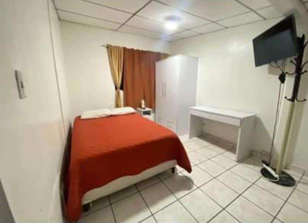 A room features a spacious bed covered with a red blanket. A wooden desk and a wardrobe are present, along with a wall-mounted television. Natural light filters through the curtains, which are complemented by a warm orange color.
