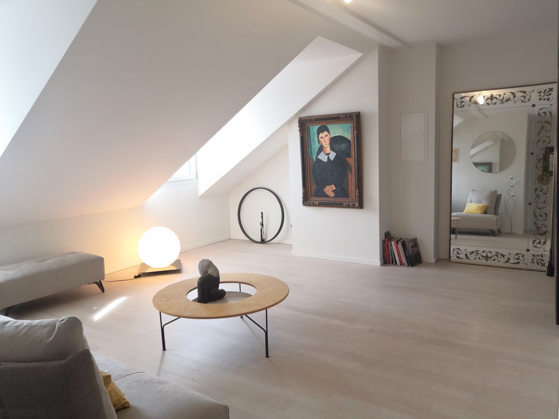 The spacious room is characterized by sloped ceilings and natural light streaming through a large window. A round coffee table is placed centrally, accompanied by a modern lamp and decorative art. A full-length mirror reflects the surrounding artwork, contributing to a serene atmosphere.