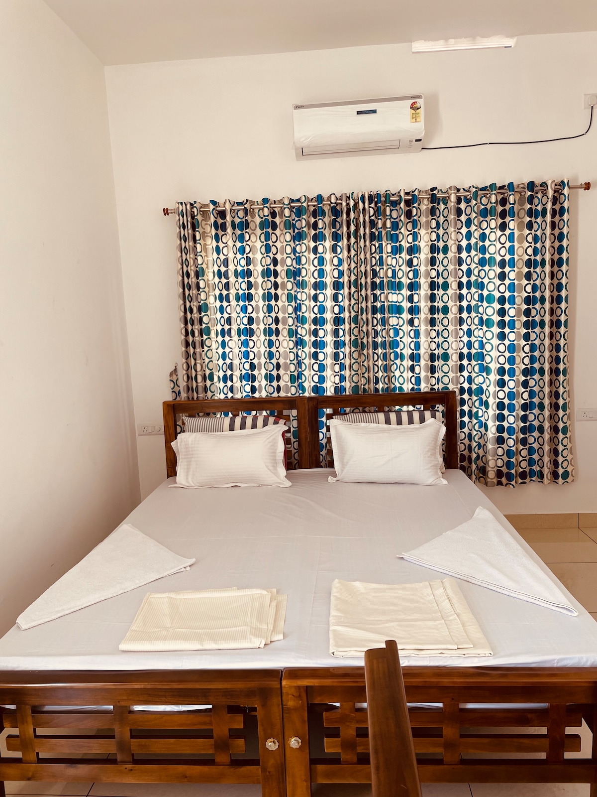 A well-lit room features a wooden bed adorned with white linens and two pillows. Blue and white patterned curtains frame a window, allowing natural light to fill the space. An air conditioning unit is mounted above, providing comfort during warm days.
