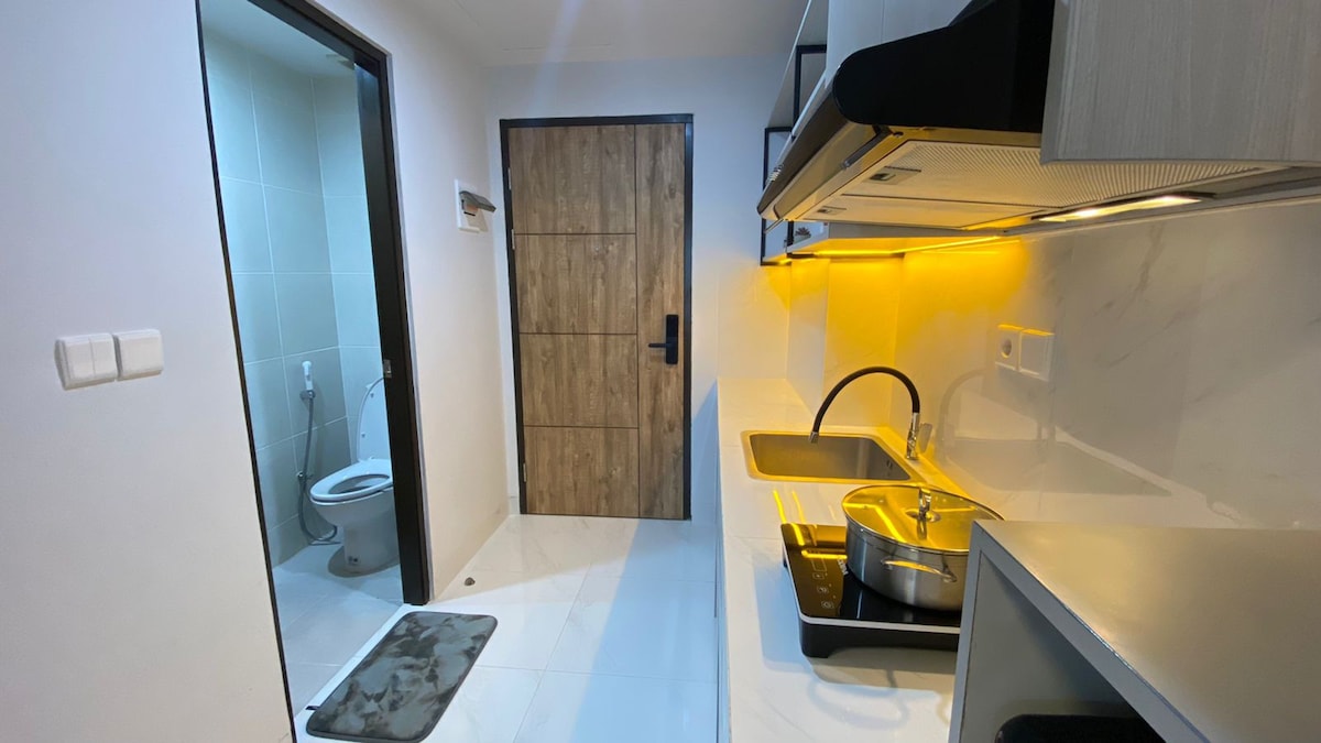 A compact kitchen area features sleek cabinetry and modern appliances. A pot sits on the stove, while soft lighting from above enhances the space. A doorway to the bathroom is visible, showcasing neutral tiles and a modern toilet.