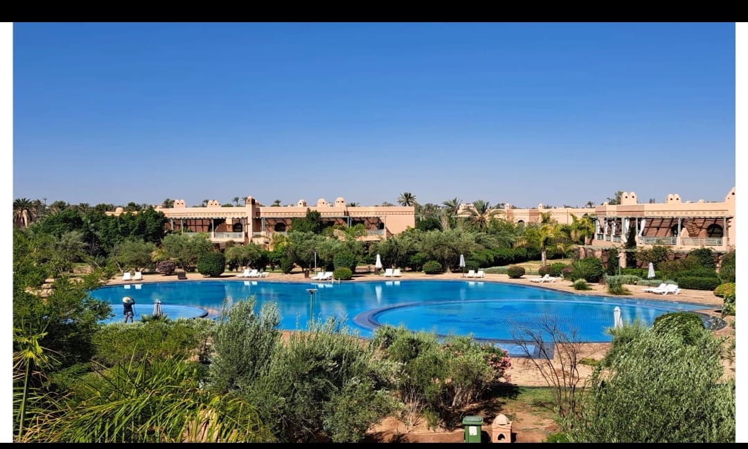 A large, inviting swimming pool is surrounded by lush greenery and well-maintained landscaping. Several lounge chairs are positioned around the pool, offering space for relaxation. The clear blue sky creates a serene atmosphere, complementing the tranquil setting.