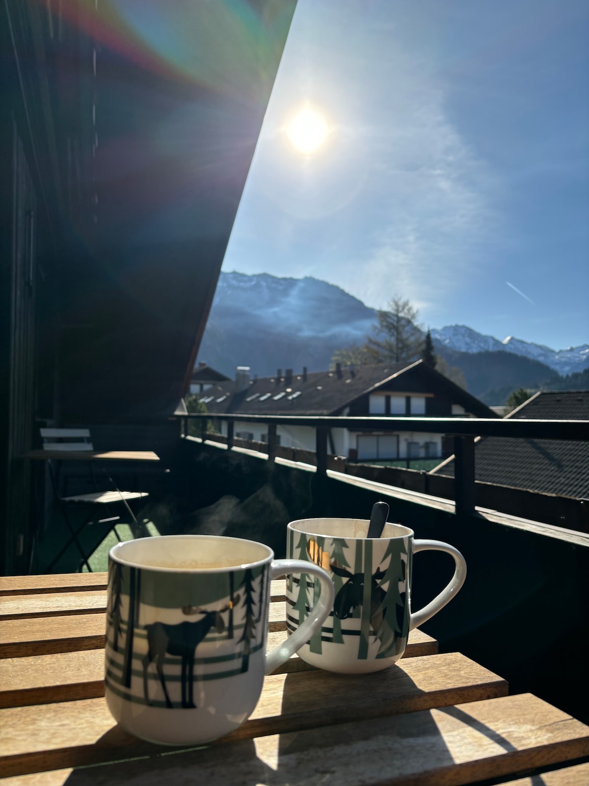 Two steaming mugs are positioned on a wooden table, with a bright sun visible in the clear sky. The background features a picturesque mountain range and nearby houses, creating a relaxing balcony atmosphere.