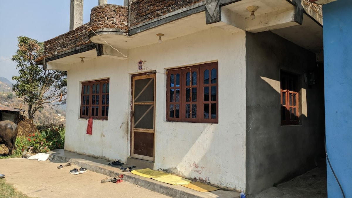 A modest building with a simple facade features multiple wooden-framed windows, allowing natural light to enter. A covered entrance is visible, and items are scattered in front of the house, including footwear and yellow mats, indicating a welcoming space for gathering.