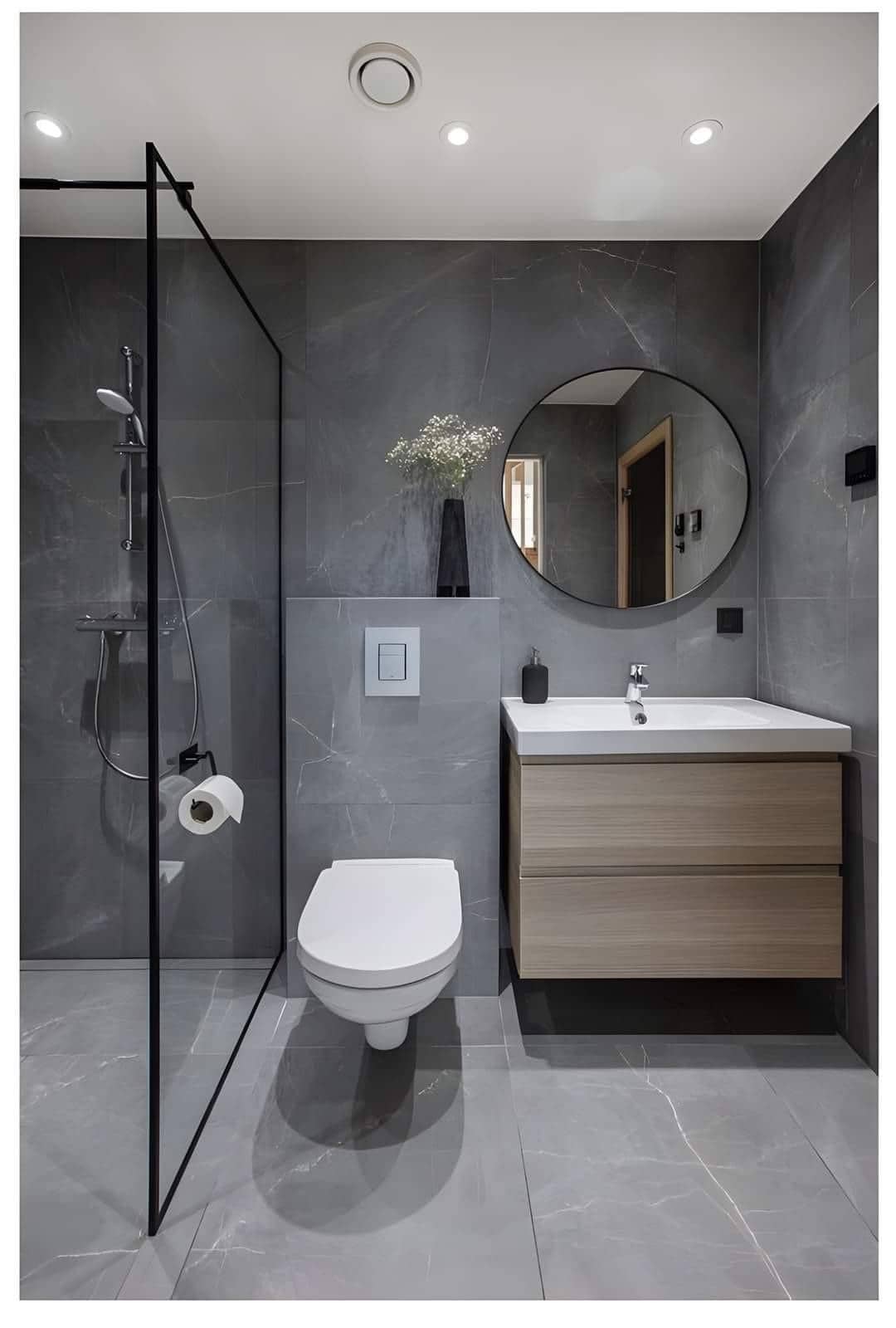 A modern bathroom features sleek dark marble walls and illuminated ceiling lights. A glass shower enclosure is present alongside a minimalist vanity with natural wood drawers. A round mirror complements the clean lines, and neutral accessories add functionality.