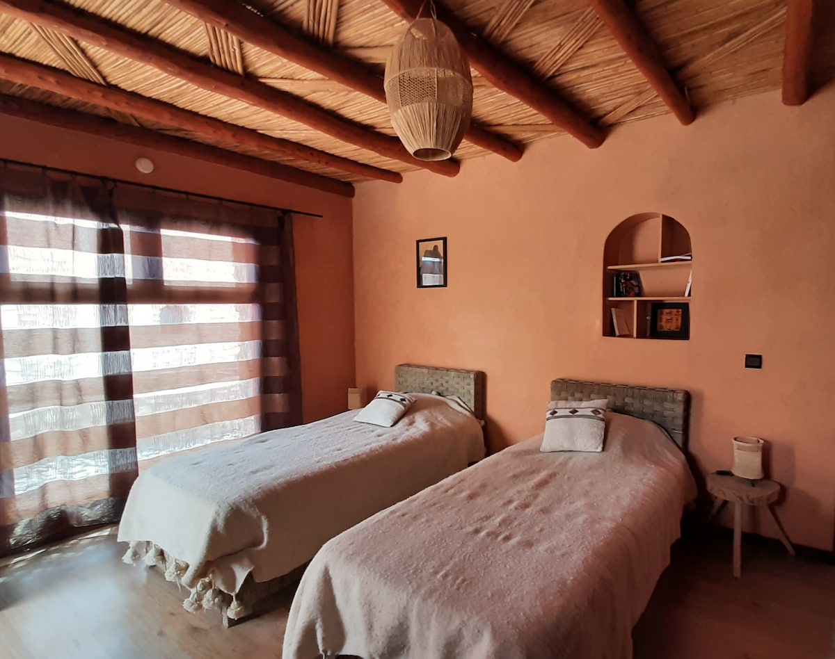 A cozy bedroom features two twin beds with soft bedding, arranged with decorative pillows. Natural light enters through a large window, adorned with sheer and opaque curtains. A wooden shelf holds decorative items, and a woven pendant light adds a rustic touch to the space.