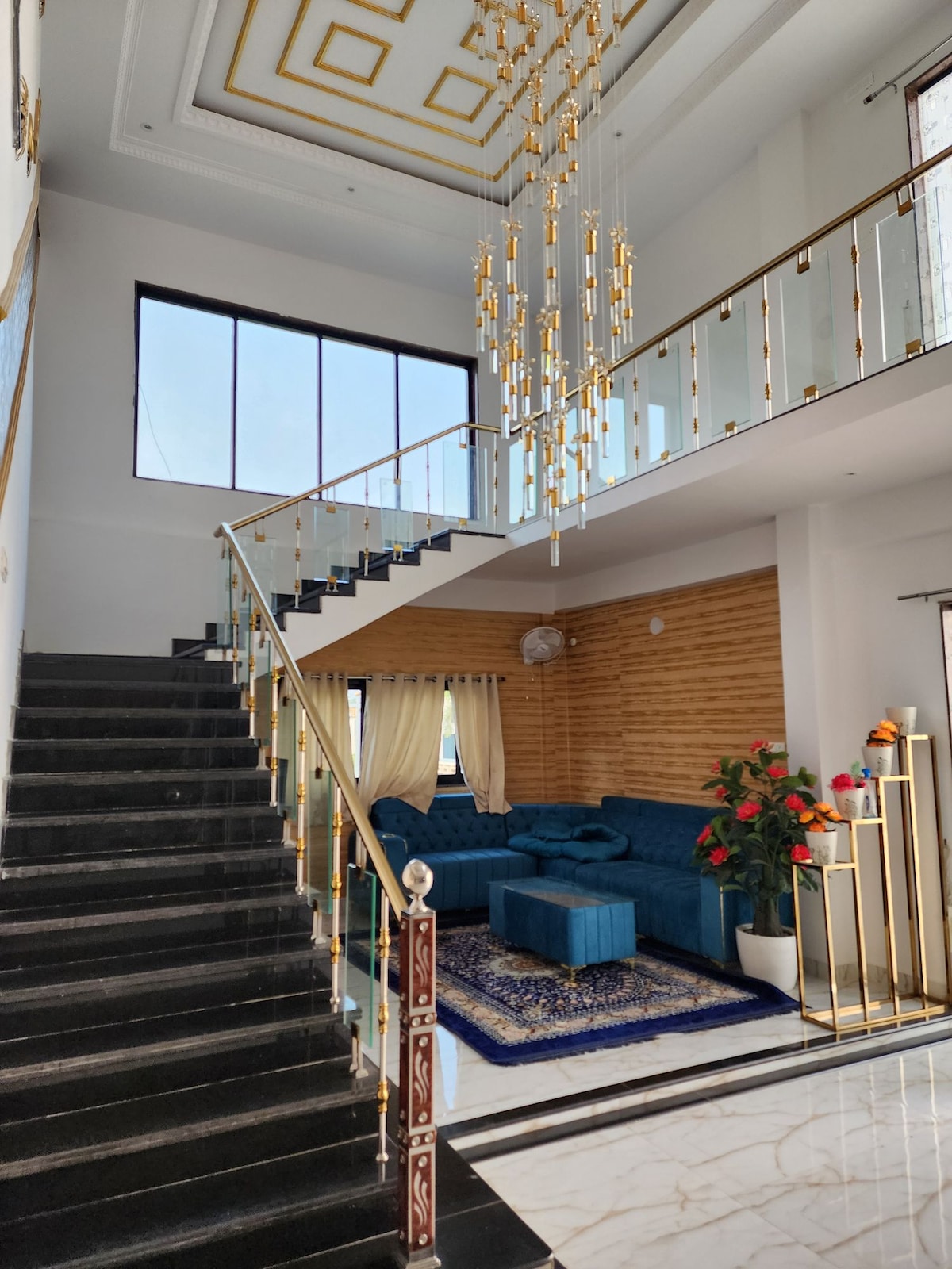 A grand entrance features an elegant staircase with glass railings, leading to an upper level. Natural light floods the space through a large window. A plush blue sofa is positioned beside a decorative plant, adding a touch of color against the neutral wall backdrop.