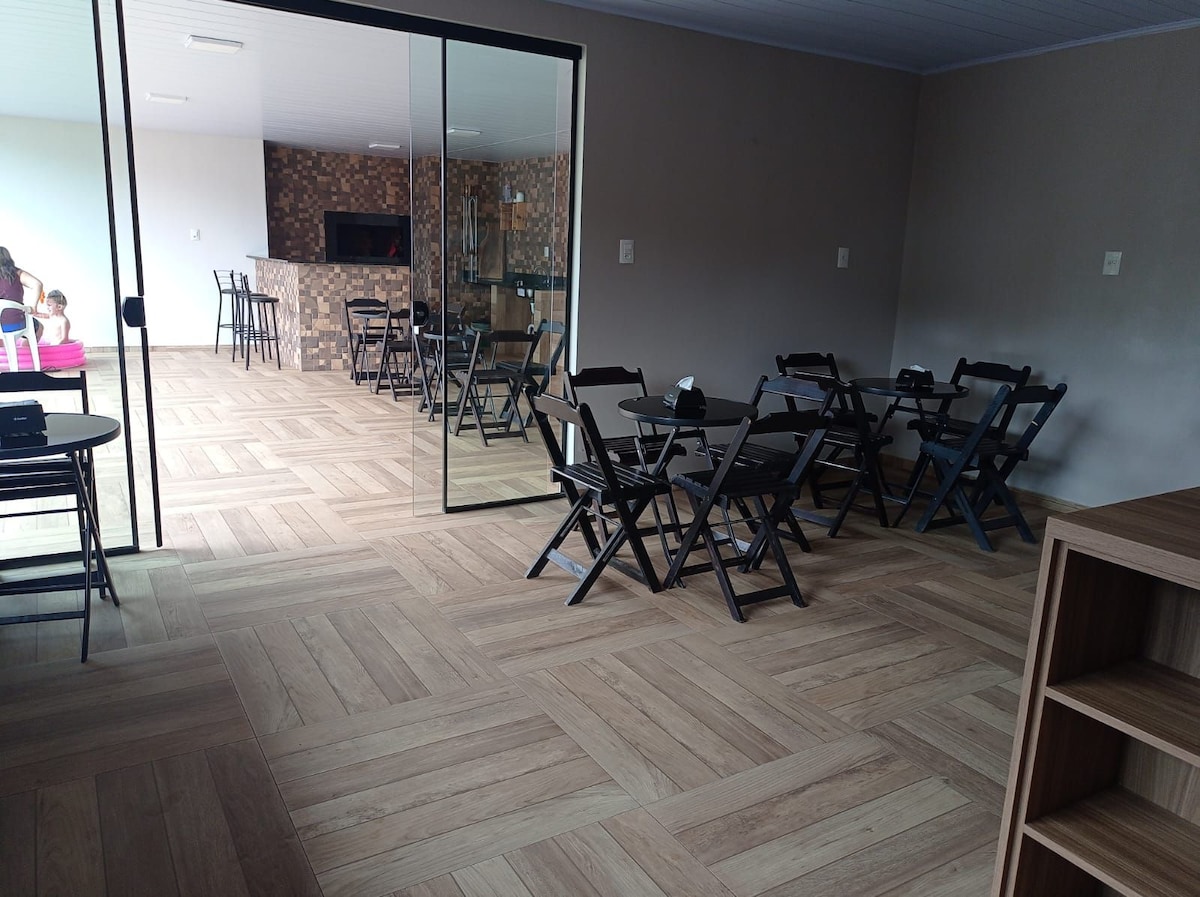 An interior space is presented, featuring a spacious area with wooden flooring. Several black folding tables and chairs are arranged throughout. Large glass doors provide access to an outdoor area, while a stone wall serves as a backdrop, contributing to a modern atmosphere.