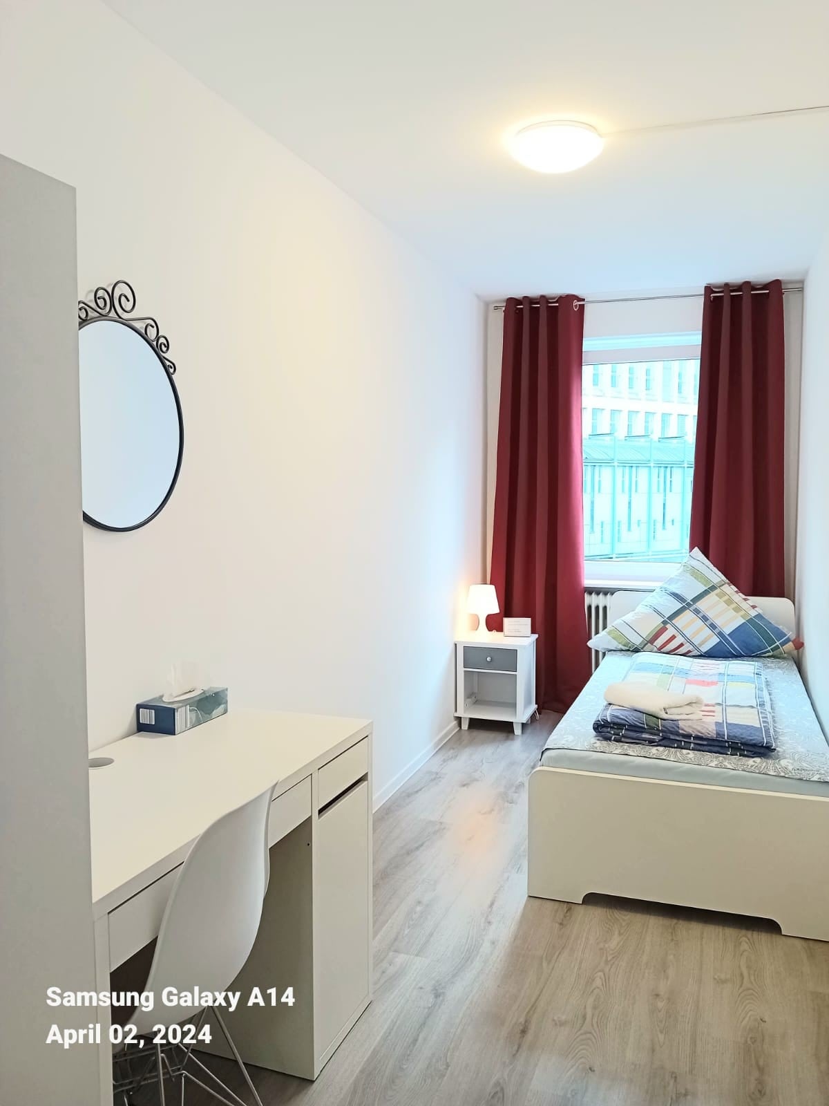 A bright and minimalistic bedroom features a single bed with a neatly made mattress, accented by a patterned throw blanket. A small desk with a chair is positioned beside the bed, while a round mirror adorns the wall. Natural light streams in through a window, with white curtains adding privacy.