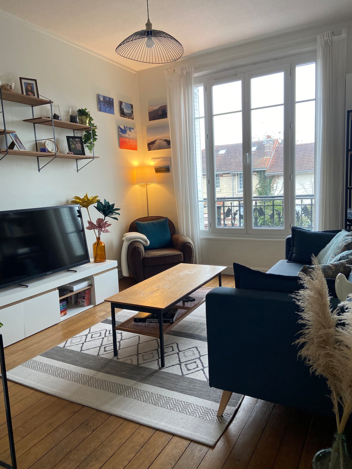 The living area features a TV cabinet and a cozy seating arrangement, including a dark blue sofa and an armchair. A wooden coffee table is positioned on a light rug, with wall shelves displaying various decorative items. Natural light filters through large windows, enhancing the room's brightness.