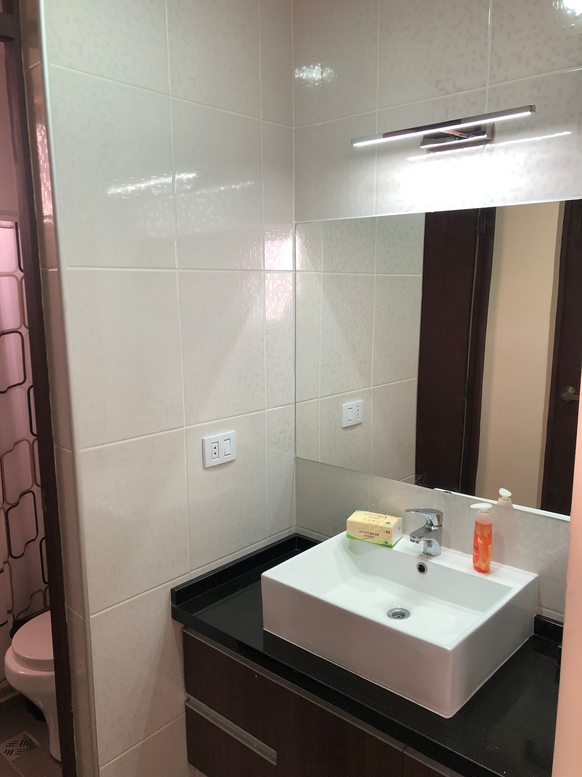 The bathroom features a modern sink with a minimalist design, positioned beneath a well-lit mirror. A neutral-toned tile wall complements the dark countertop. Visible in the background, a partially open door leads to a shower area, enhancing the bathroom's functionality.