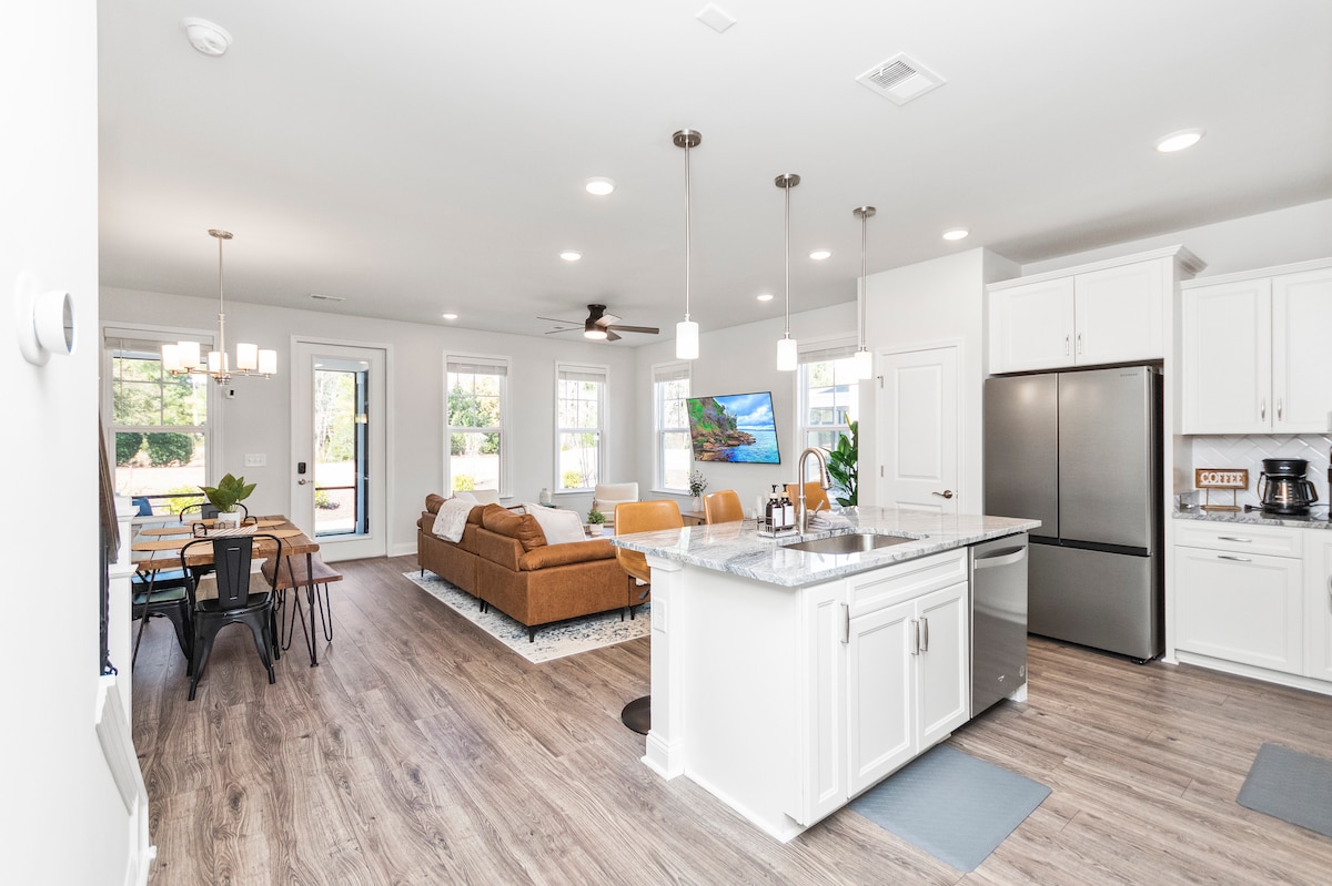An open-concept living area showcases a modern kitchen with stainless steel appliances and a spacious countertop. A cozy brown sofa is positioned in the living room, with large windows providing natural light. A dining area with a table is visible, offering seating for six.