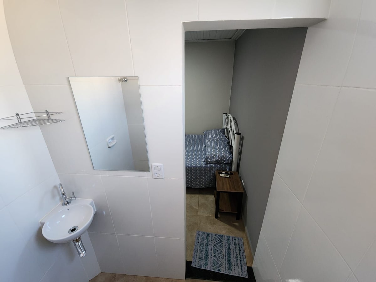 A compact room is visible, featuring a small bed adorned with blue and white bedding. A mirror reflects part of the space. A wall-mounted sink is positioned in the foreground, and a bedside table sits next to the bed, providing additional functionality.