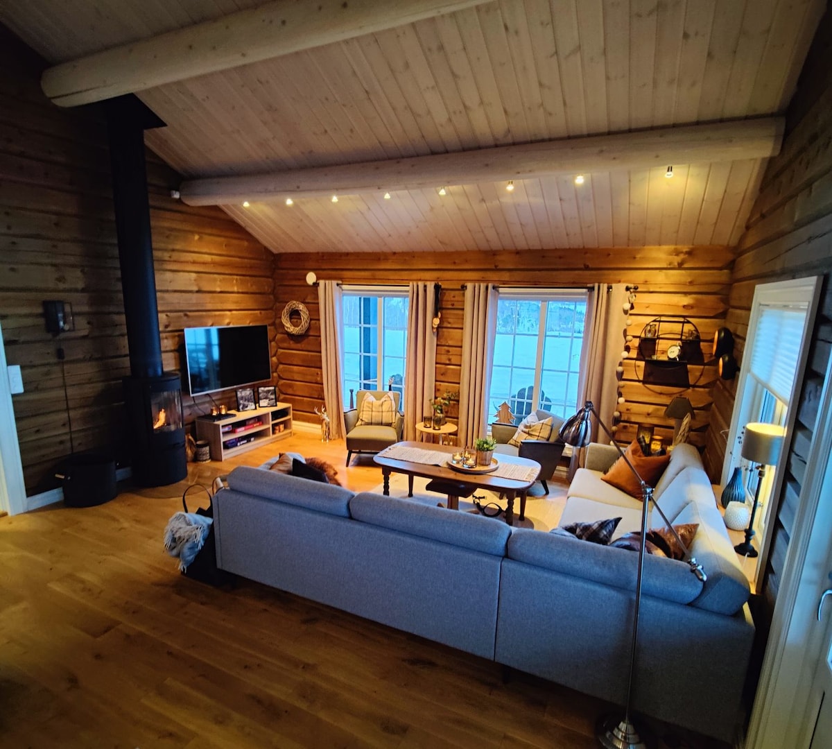 A spacious living area features an inviting sectional sofa with a soft design. Large windows allow natural light to fill the room, highlighting wooden walls and ceiling. A modern television is mounted on the wall, and a cozy fireplace adds warmth to the ambiance.