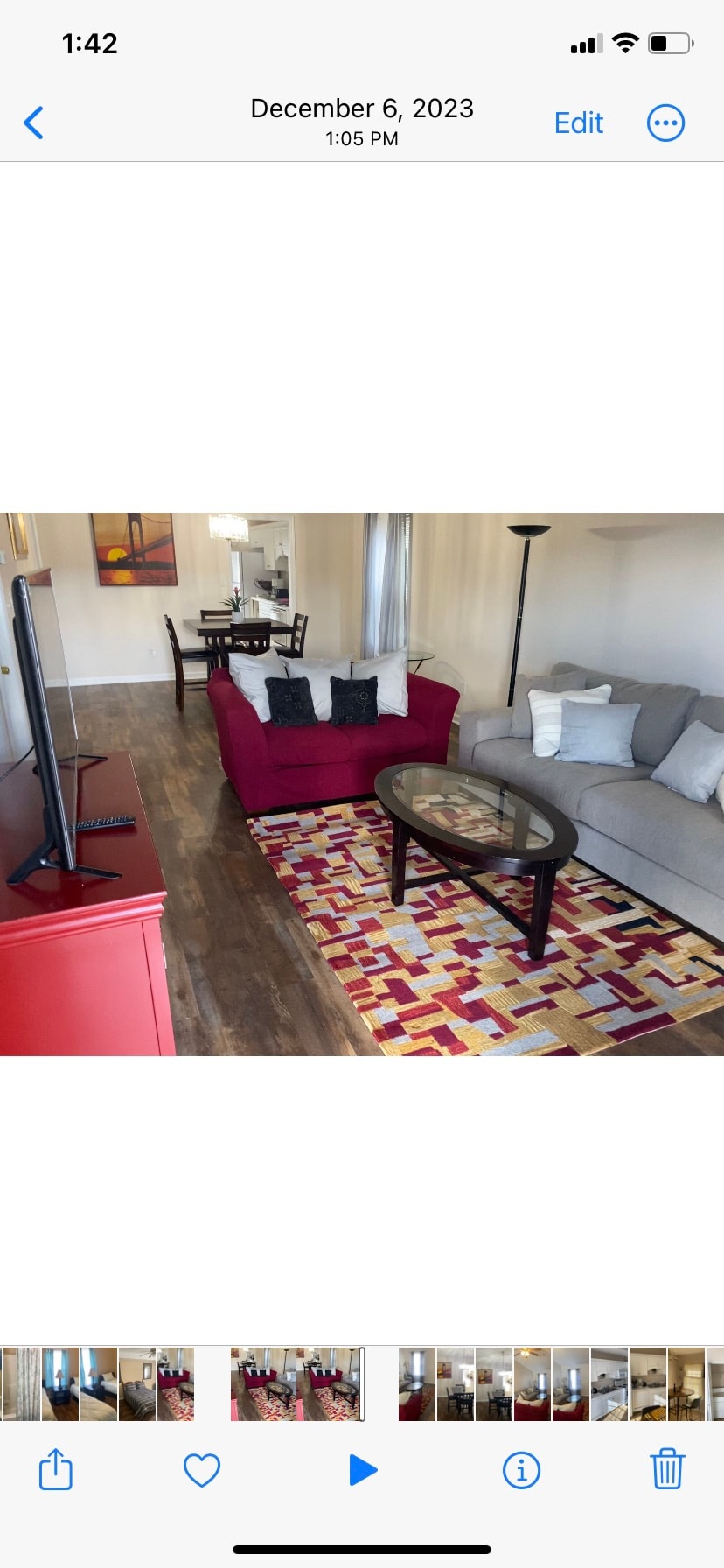The living area features a mix of cozy seating with a red couch and light gray sectional. A low coffee table sits atop a colorful area rug. A television is positioned on a red entertainment unit, with a dining table visible in the background.