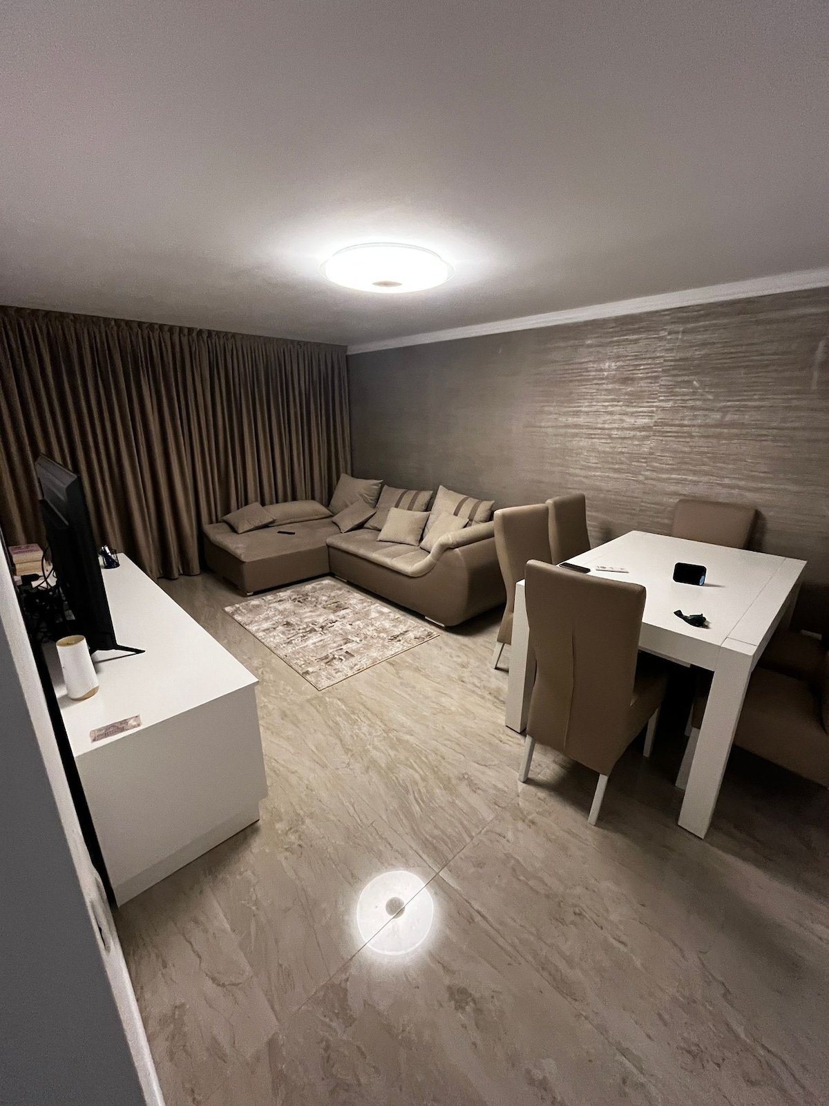 A light-filled living area features a comfortable sectional sofa positioned adjacent to a sleek white dining table surrounded by four upholstered chairs. A soft area rug adds warmth, while neutral-toned curtains frame the space, creating a serene retreat.