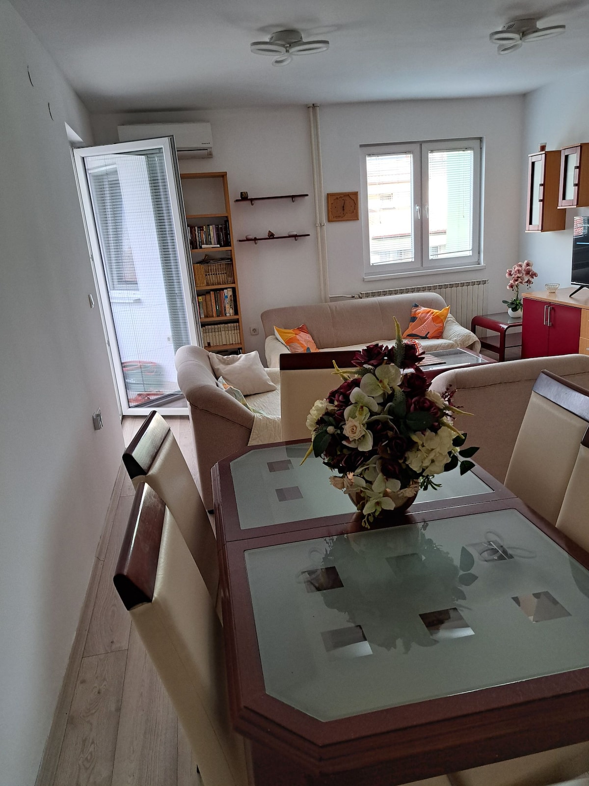 A cozy living area features a light-colored sofa with decorative cushions and a wooden coffee table, which is surrounded by dining chairs. A small bookshelf is visible on the wall, and light enters through the glass doors that open to a balcony.
