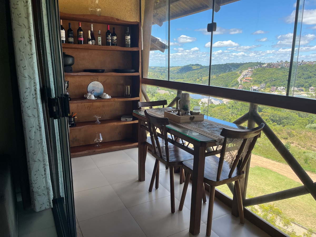 A cozy balcony is presented with a wooden dining table surrounded by four chairs. A shelf displays an assortment of bottles, complementing the panoramic view of lush green hills and a clear blue sky.