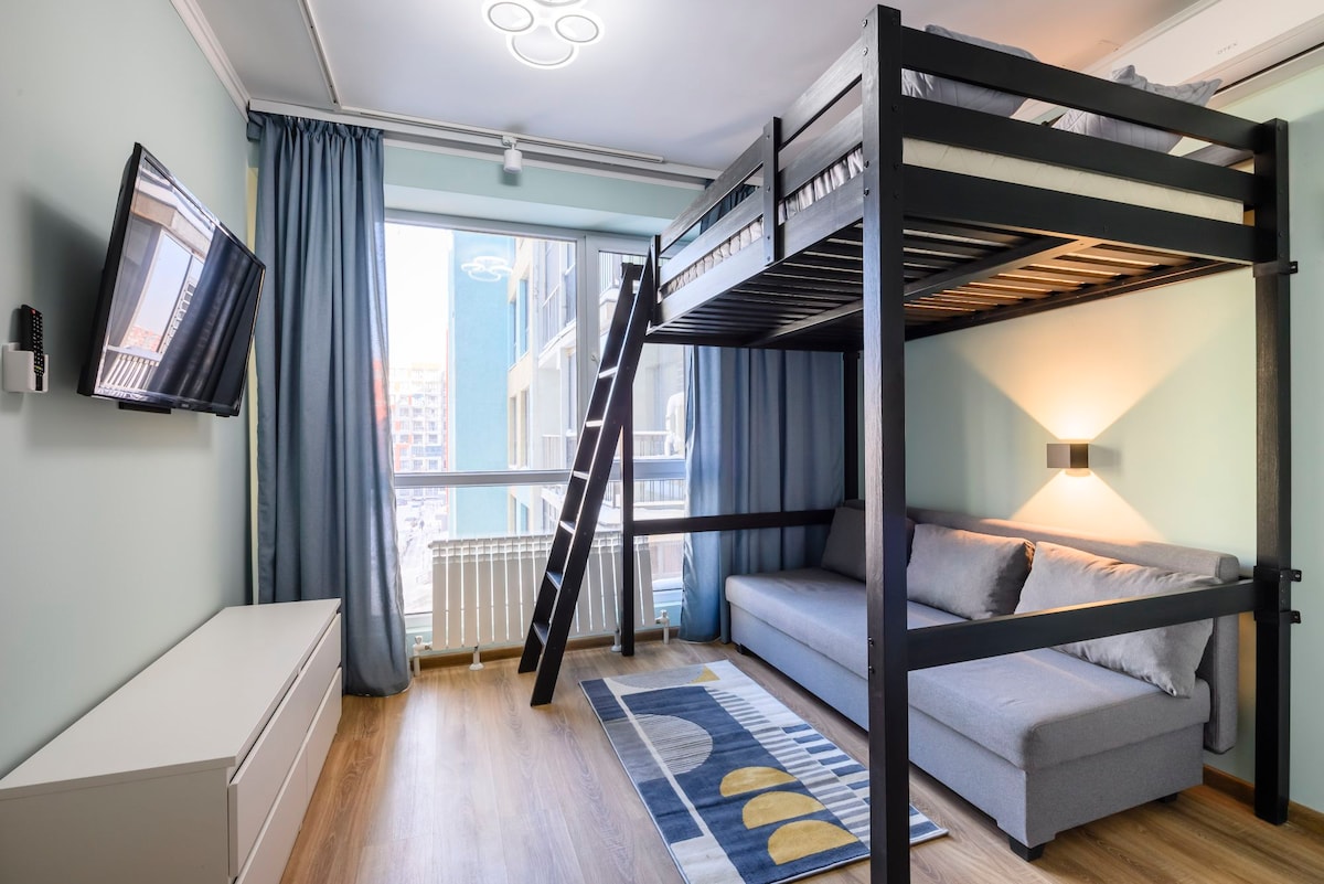 A lofted bed is positioned above a compact sofa, providing a functional use of space. A bright window offers natural light, while soft curtains frame the view. A modern television is mounted on the wall, and a rug adds comfort to the wooden floor.
