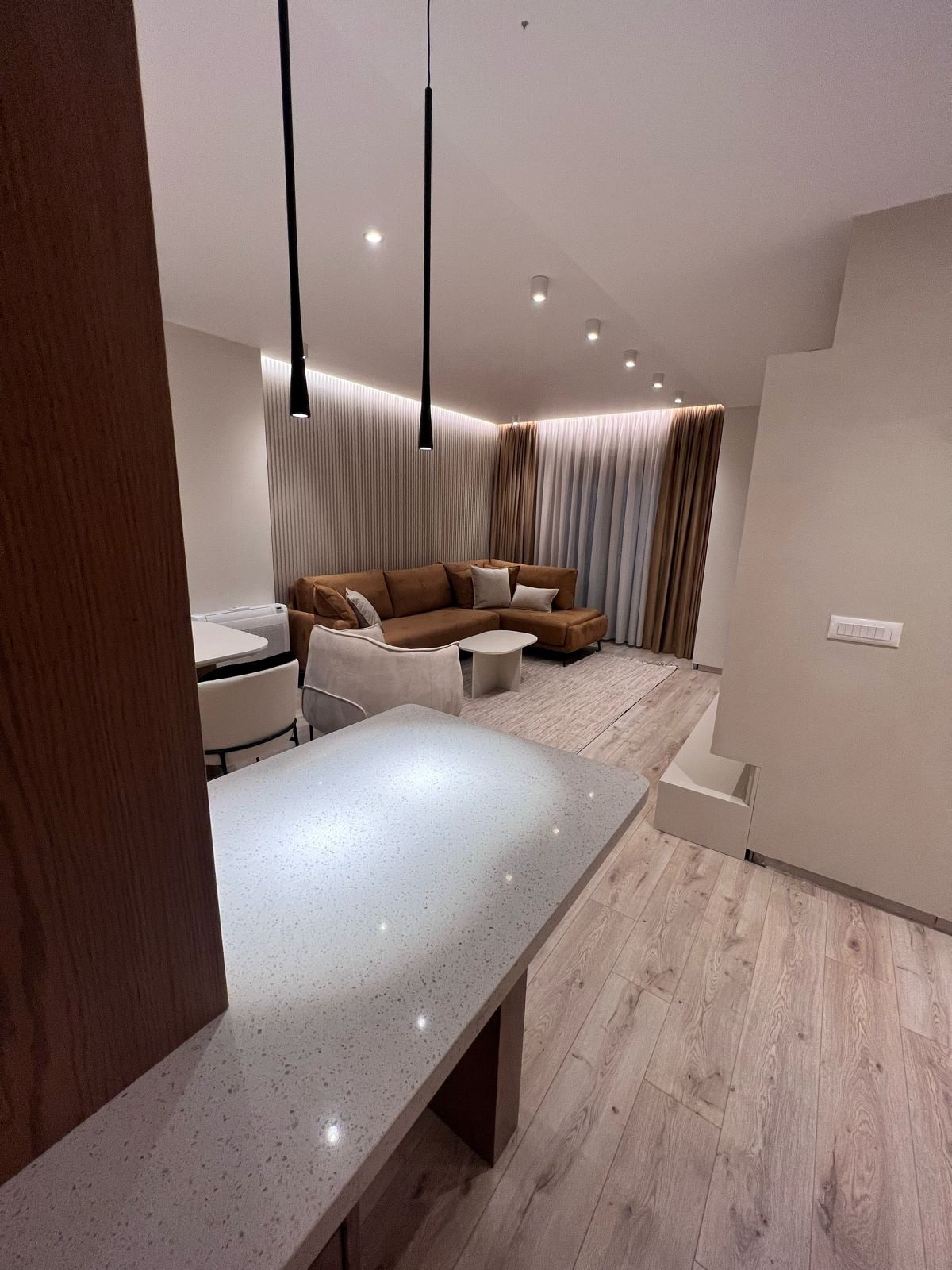 The spacious living area is presented with a comfortable couch and a small coffee table. Soft lighting from recessed fixtures enhances the atmosphere. A chic dining setup is visible, featuring a table surrounded by light-colored chairs. Warm tones dominate the decor, complemented by floor-to-ceiling curtains.