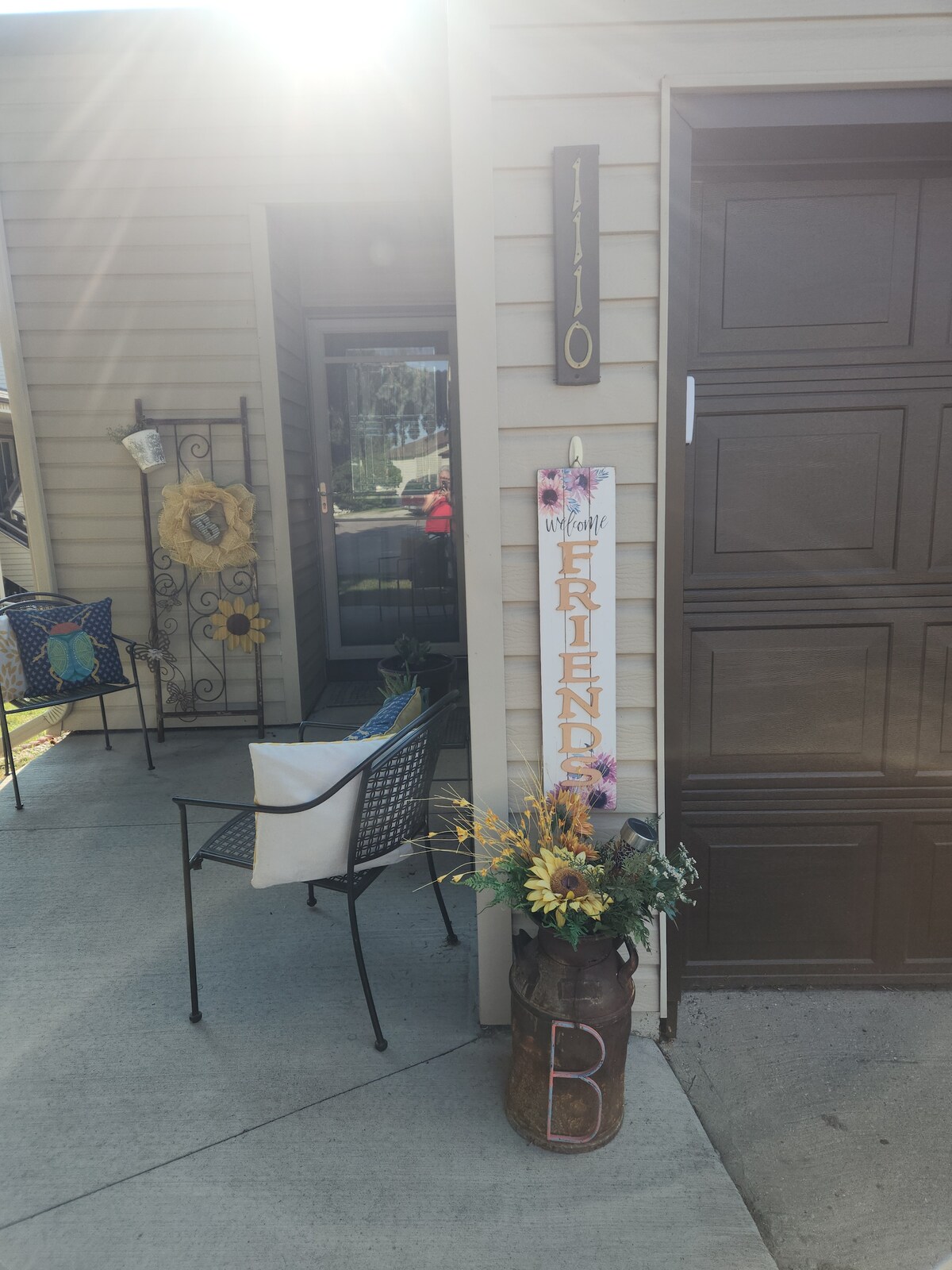A welcoming entrance is featured, showcasing a light-colored facade and a dark garage door. A decorative sign reading 'Friends' is positioned next to a rustic planter filled with yellow flowers. A chair sits nearby, providing a cozy spot for guests.