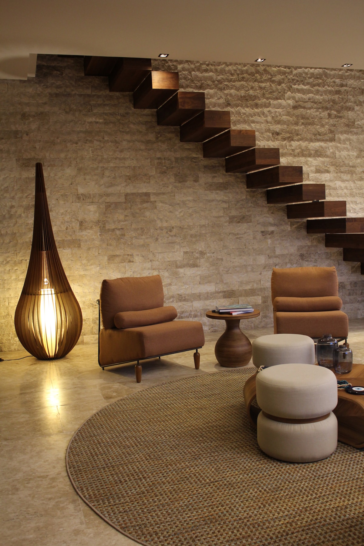 A modern living area is showcased, featuring two stylish chairs and a round coffee table. A sculptural light fixture provides soft illumination beside a unique staircase made of wood, enhancing the space's elegant design. A round rug adds warmth to the polished floor.