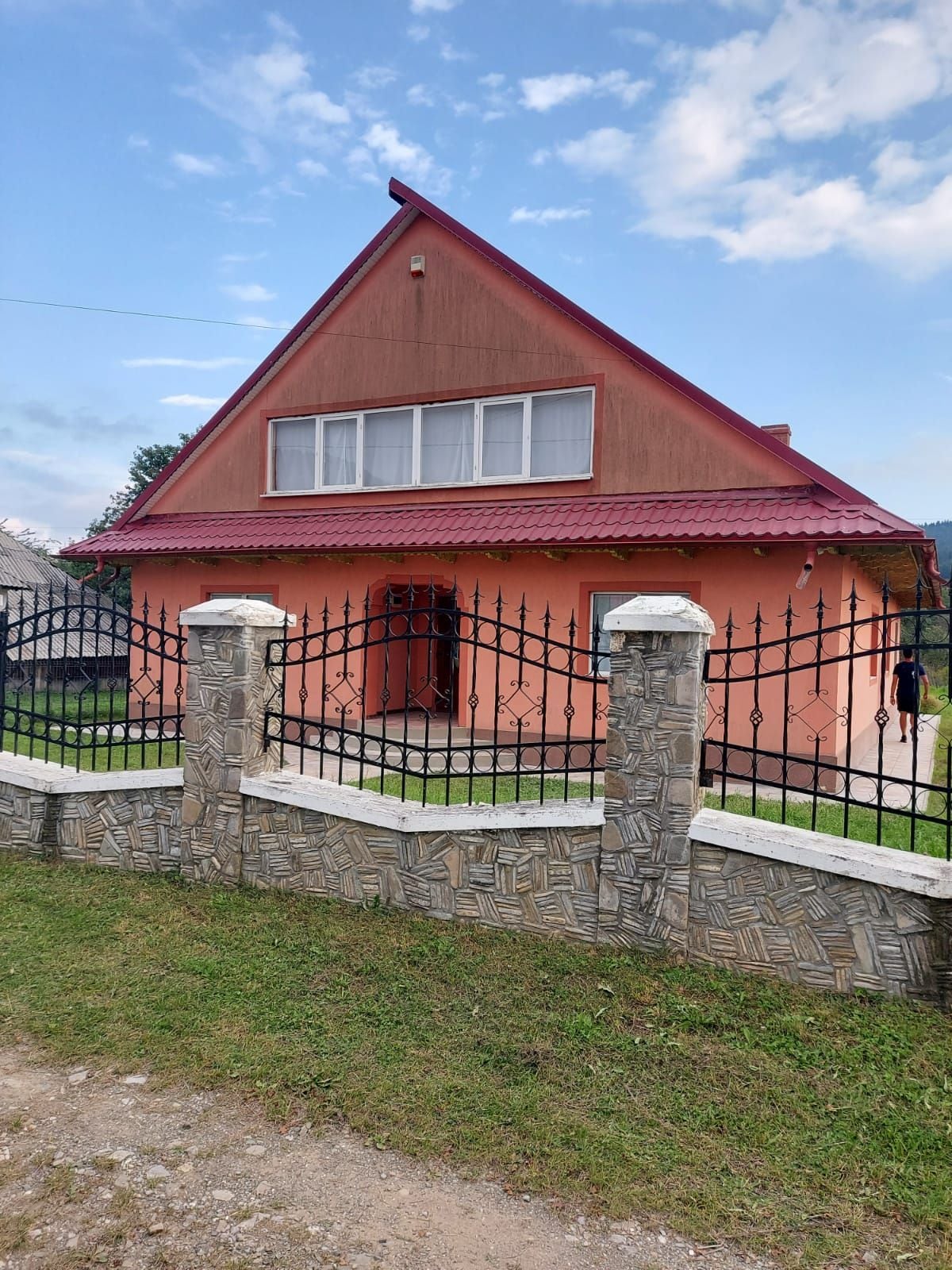 A pink house with a triangular roof is enclosed by an ornate black iron gate. The facade features large windows on the upper level, and greenery surrounds the property, providing a serene environment.