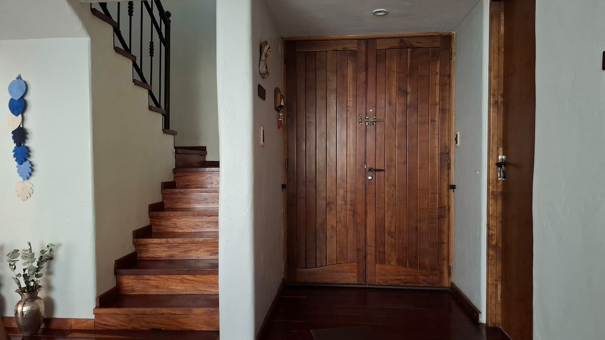 A welcoming entrance features a pair of solid wooden doors, with decorative wall accents visible nearby. A staircase with a wooden railing leads to an upper level, while a potted plant adds a touch of greenery to the entryway.