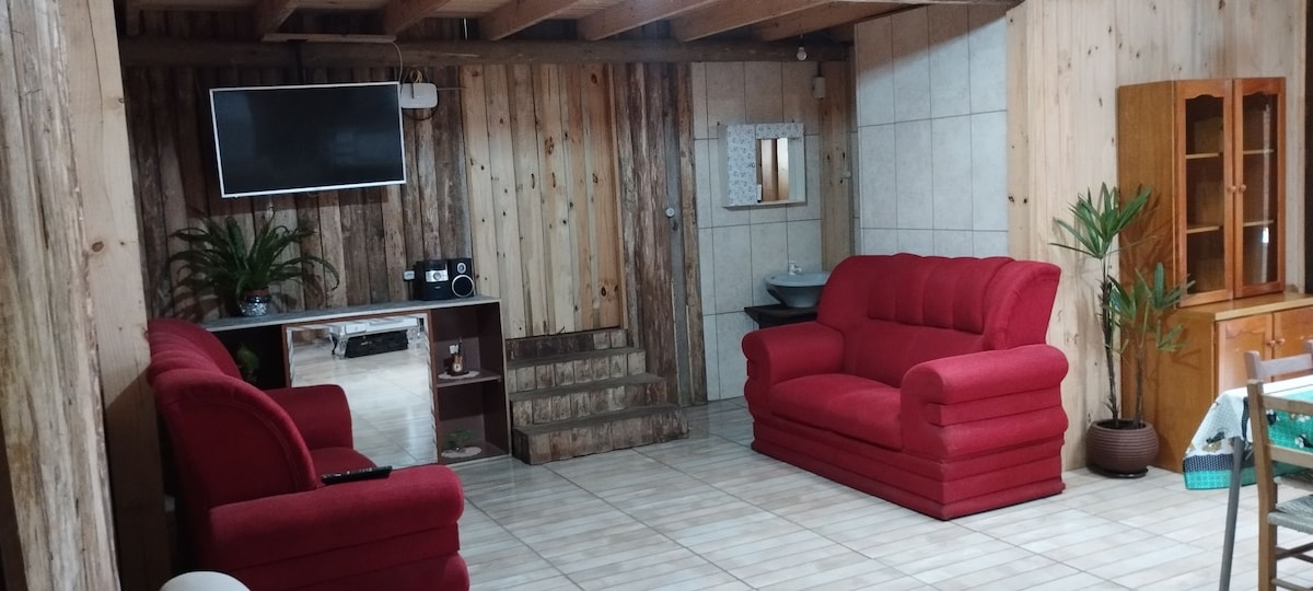 A spacious living area features wooden walls and ceilings, complemented by tiled flooring. Two red sofas offer comfortable seating, while a wall-mounted TV is positioned above a decorative console. A small step leads to a wash area, and a plant adds a touch of greenery.