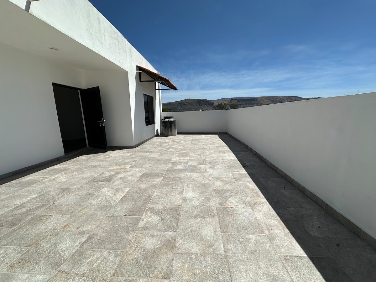 A spacious outdoor terrace features a smooth stone floor and is bordered by white walls. Breathtaking views of the surrounding landscape are visible, with clear blue skies overhead, providing an open and airy atmosphere.