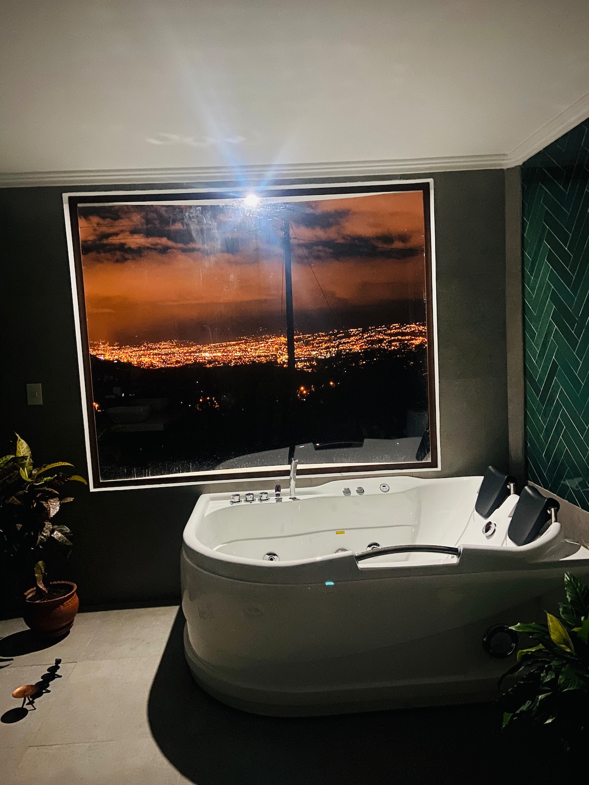 A modern bathroom features a spacious jacuzzi positioned beside a large window. The window offers a panoramic view of the city illuminated at night, while a small plant in a pot adds a touch of greenery to the serene space.