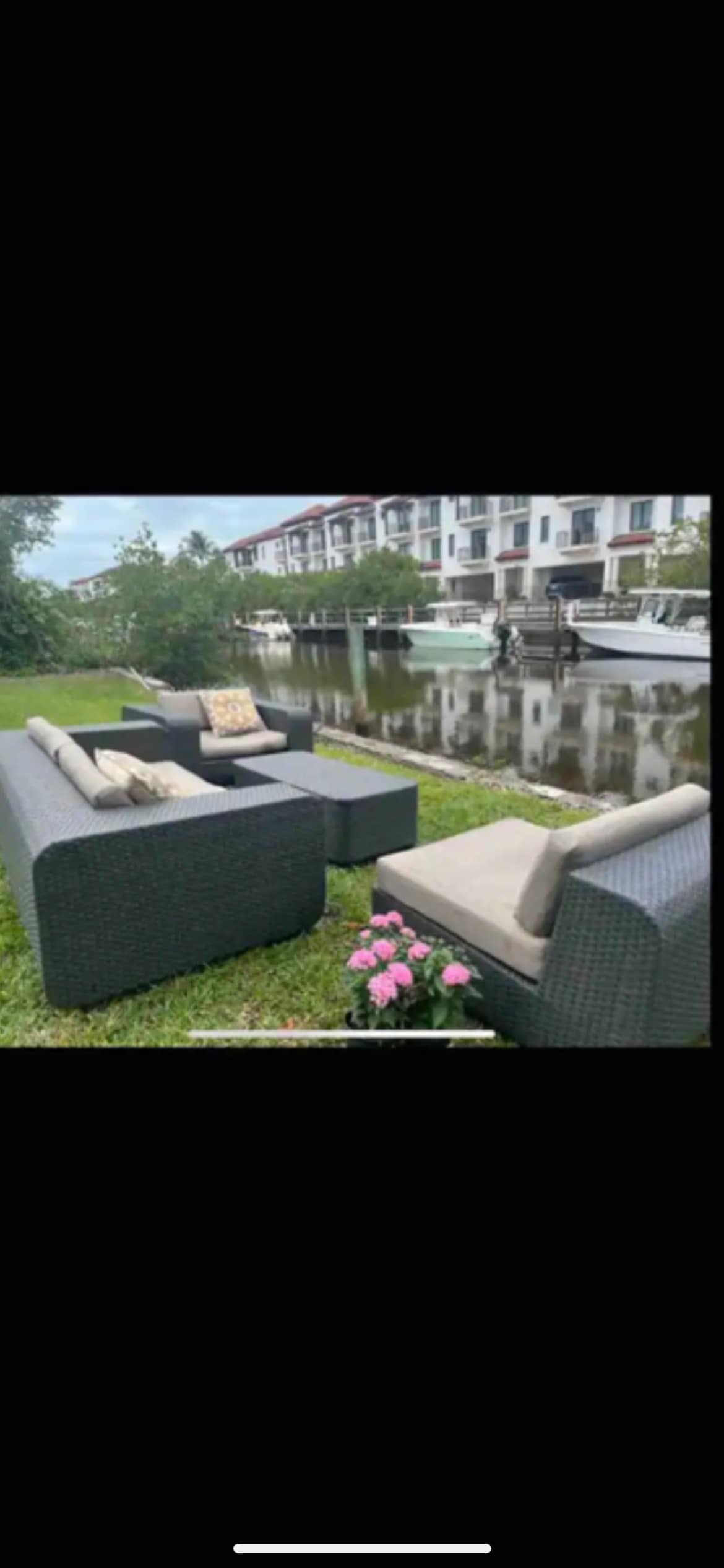 An outdoor seating area is showcased along a calm waterway, featuring a contemporary gray wicker sectional sofa with plush cushions. A table complements the seating arrangement, with colorful flowers adding a natural touch nearby. The backdrop includes a low-rise building and moored boats.