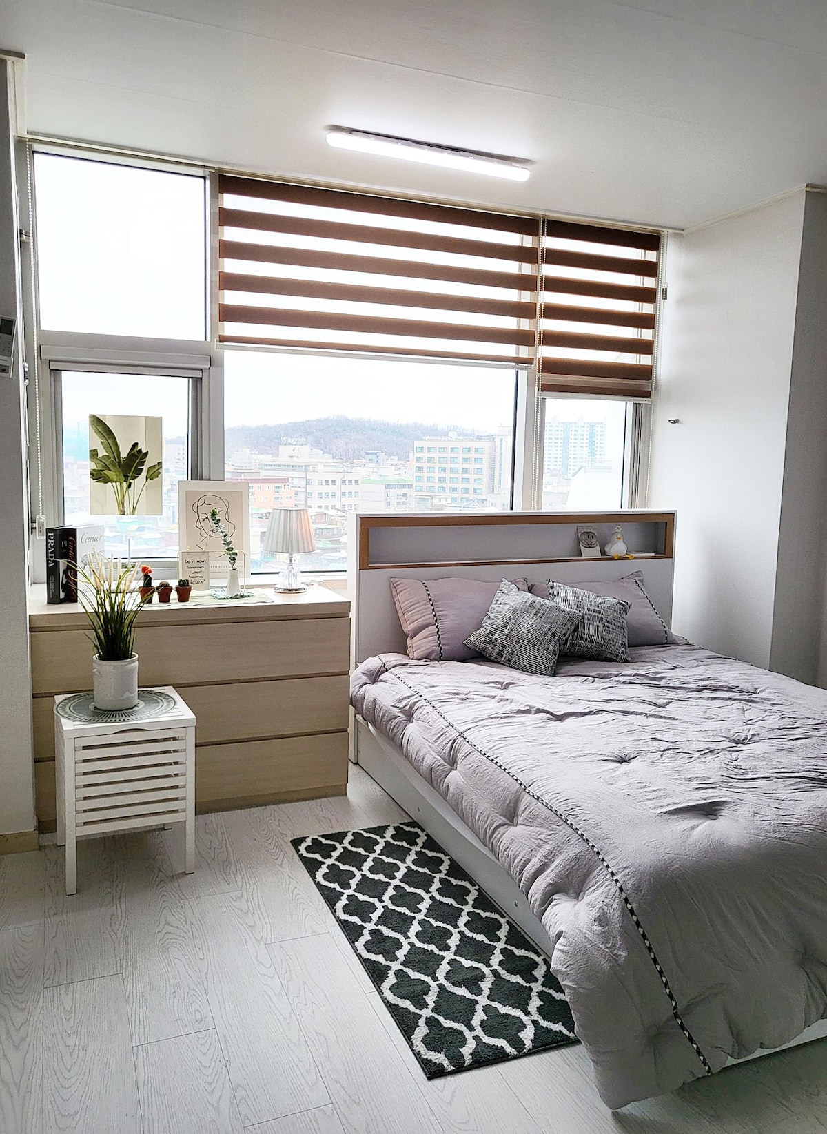 A cozy bedroom features a spacious bed dressed in soft linens and decorative pillows. Natural light streams through large windows adorned with dual-tone blinds. A bedside table holds decorative items, and a stylish rug lies on the light-colored floor.