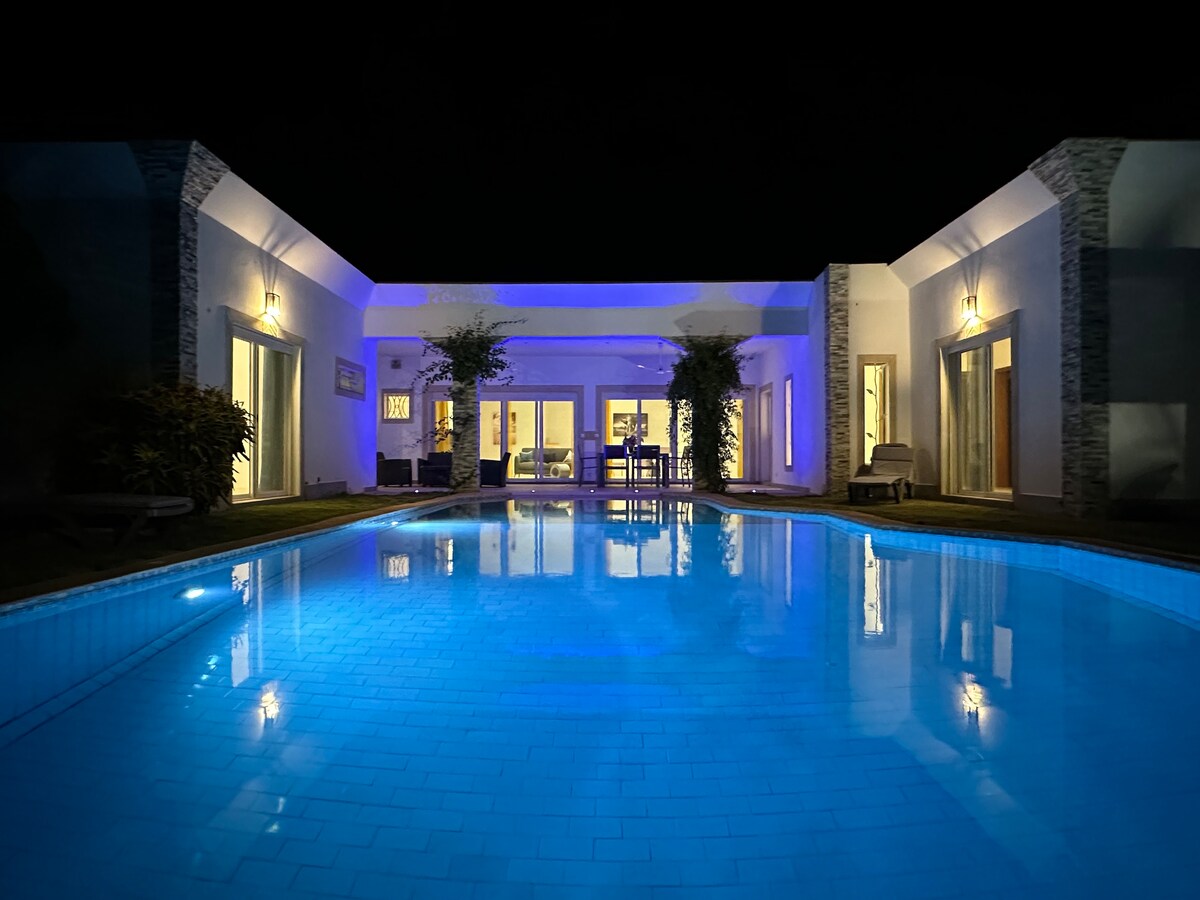 The villa is illuminated at night, showcasing a large pool with vibrant blue lighting reflecting on the water's surface. Glass doors provide a view of the spacious interior, while lounge chairs are positioned around the pool, creating an inviting outdoor area.