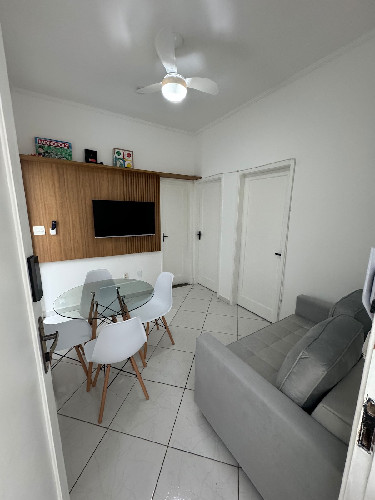 A light-filled living area features a gray sofa and a glass dining table surrounded by white chairs. A wooden wall panel supports a flat-screen TV, while two doors lead to adjacent rooms. A ceiling fan adds functionality to the space.