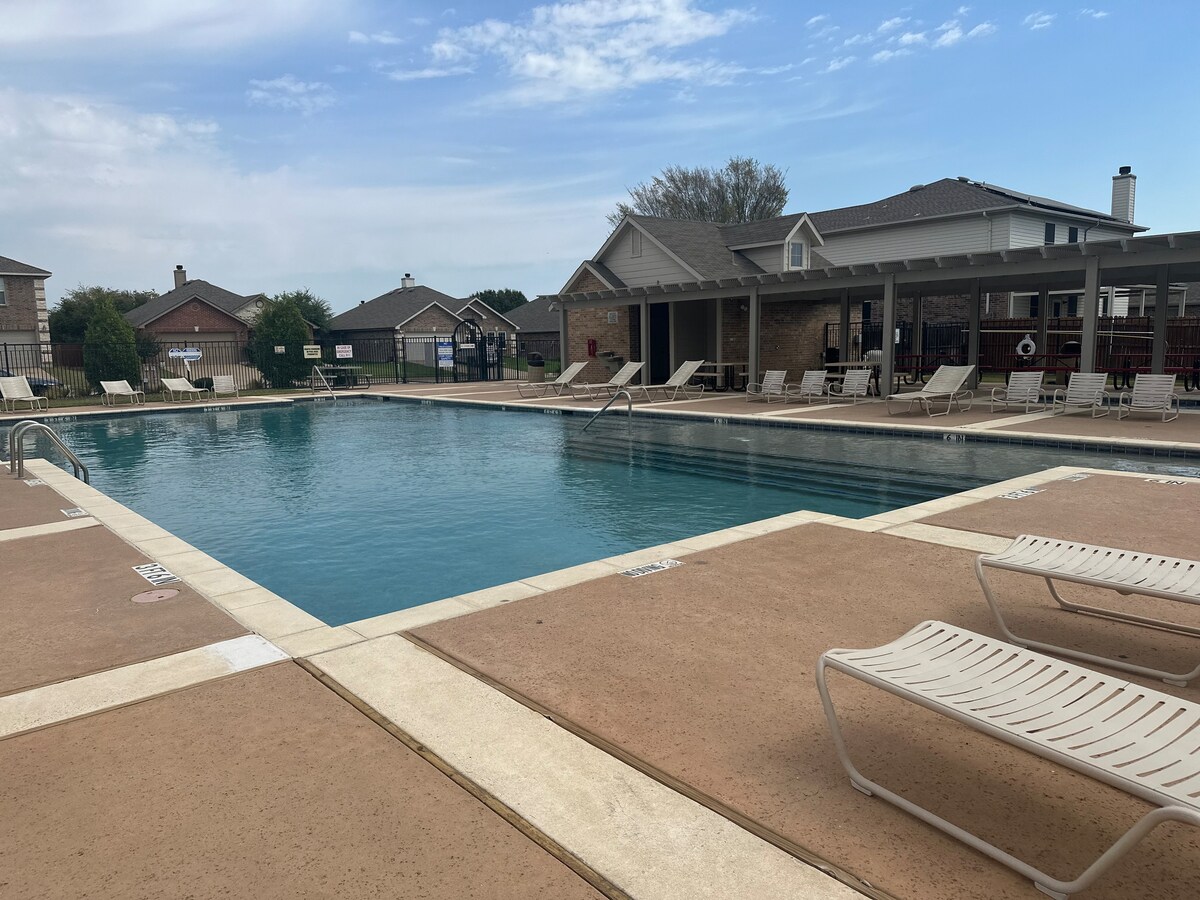 The community pool area is shown, featuring a large swimming pool surrounded by a spacious deck. Several lounge chairs are arranged neatly around the pool. A building with seating and shade is visible in the background, with residential homes located beyond.