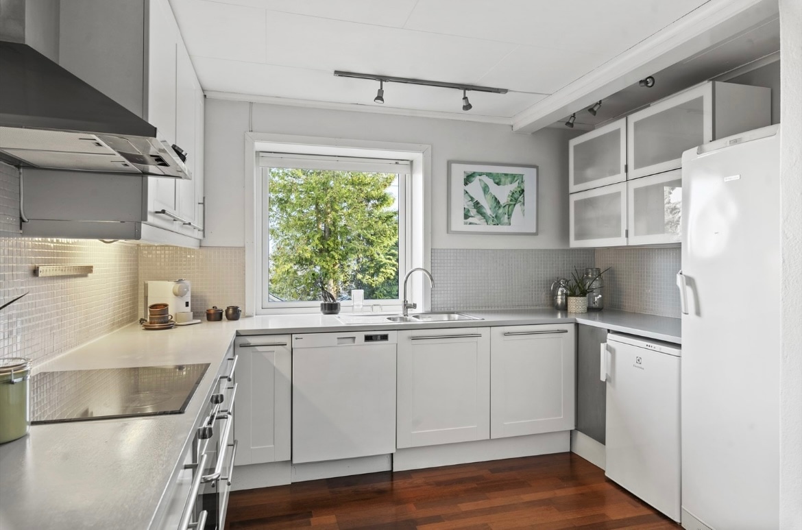 A modern kitchen features light grey cabinetry and a functional layout. Stainless steel appliances, including a refrigerator and dishwasher, are integrated seamlessly. A large window allows natural light to brighten the workspace, with a view of greenery visible outside.