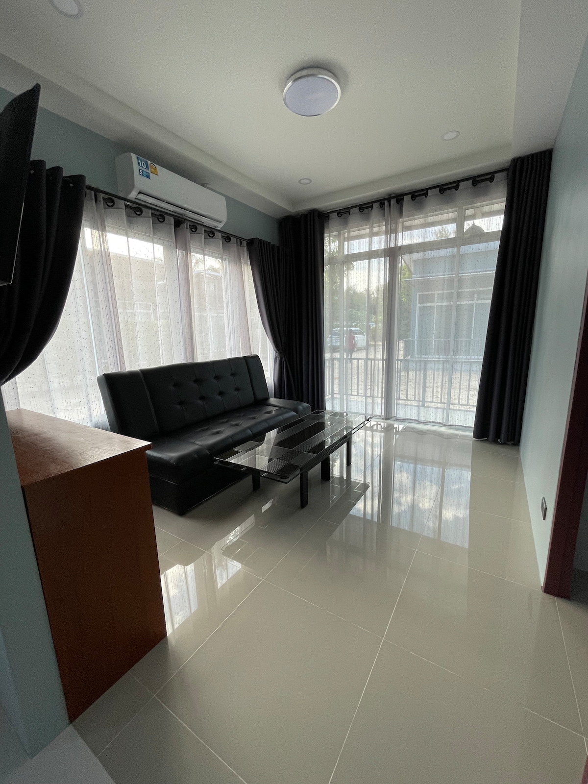A modern living area is featured with a sleek black sofa positioned across from a glass coffee table. Natural light filters through sheer curtains, illuminating the shiny tiled floor. Dark curtains frame the window, creating a balanced contrast in the space.