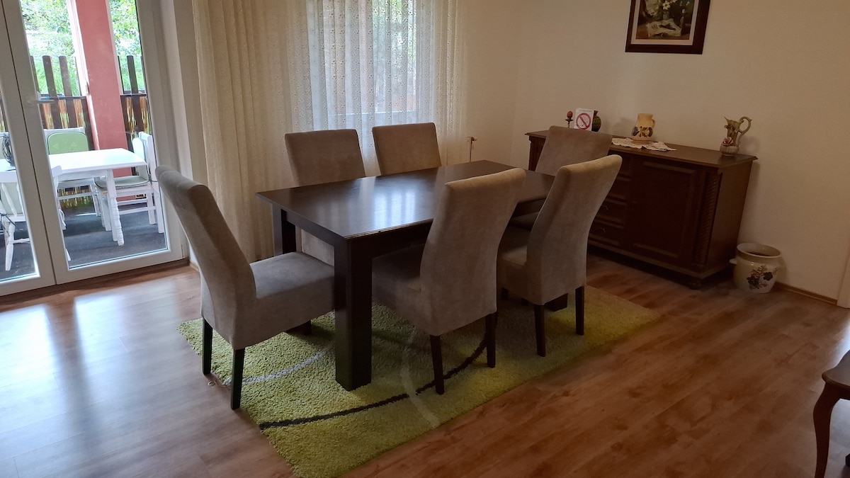 A dining area is presented with a rectangular table surrounded by six upholstered chairs. A plush area rug adds warmth beneath the table. Natural light filters through sheer curtains, illuminating the space, which is complemented by a wooden sideboard and a view of the outdoor patio.