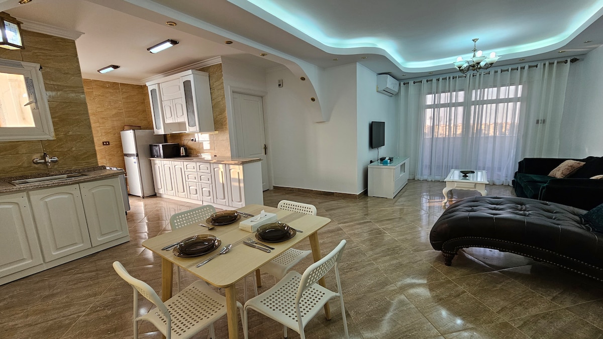 An open-plan living area features a modern kitchen equipped with white cabinetry and stainless-steel appliances. A dining table with four chairs is positioned near a large window, allowing natural light to fill the space. A comfortable sofa and a side table enhance the inviting atmosphere.