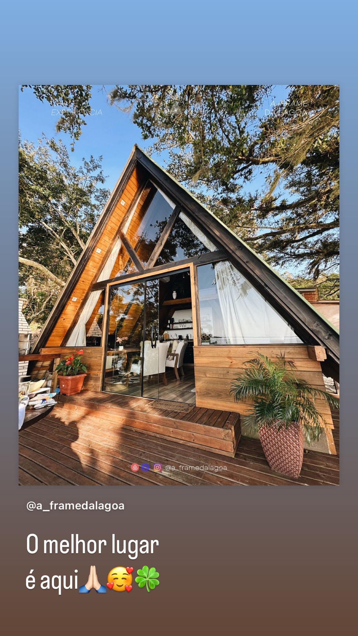 The A-frame cabin features large glass windows that allow natural light to flood the interior. A wooden deck extends from the entrance, surrounded by lush greenery. The structure's design integrates traditional and modern elements, highlighting the connection to nature.