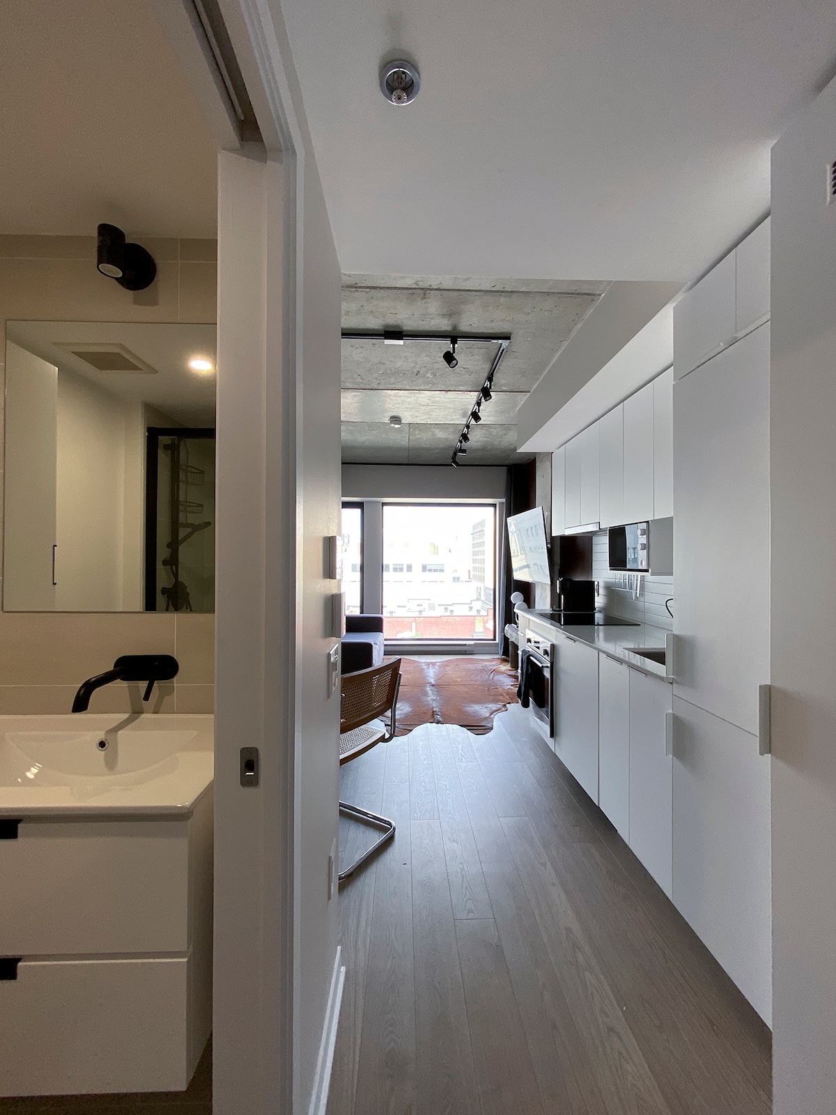 A modern hallway connects a bathroom and a kitchen area, featuring sleek cabinetry and stainless steel appliances. Natural light enters through a large glass door at the end of the hallway, leading to an outdoor view. A cozy chair is positioned nearby, adding comfort to the space.
